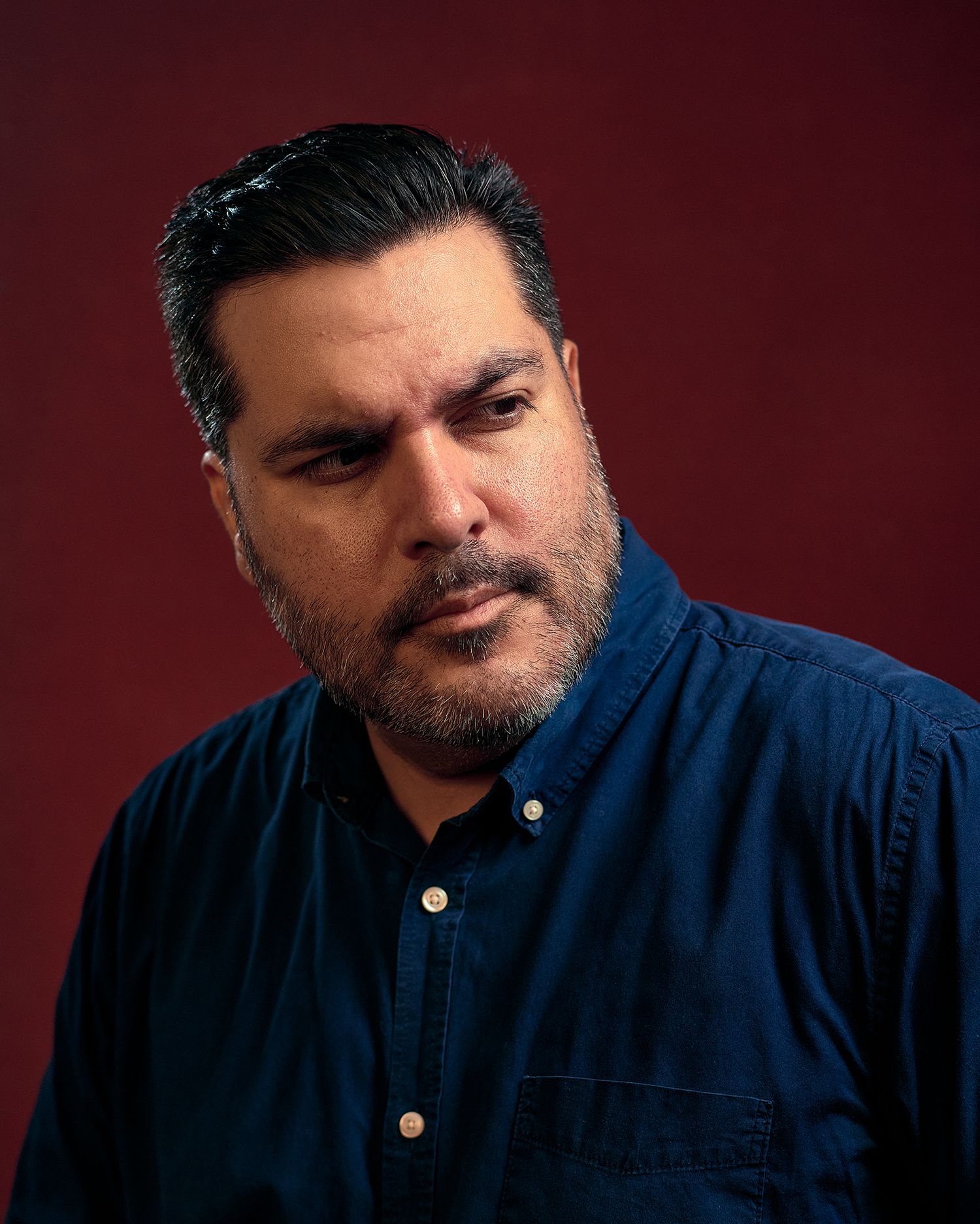 A serious man with dark hair and a beard wearing a blue denim shirt poses against a dark red background.