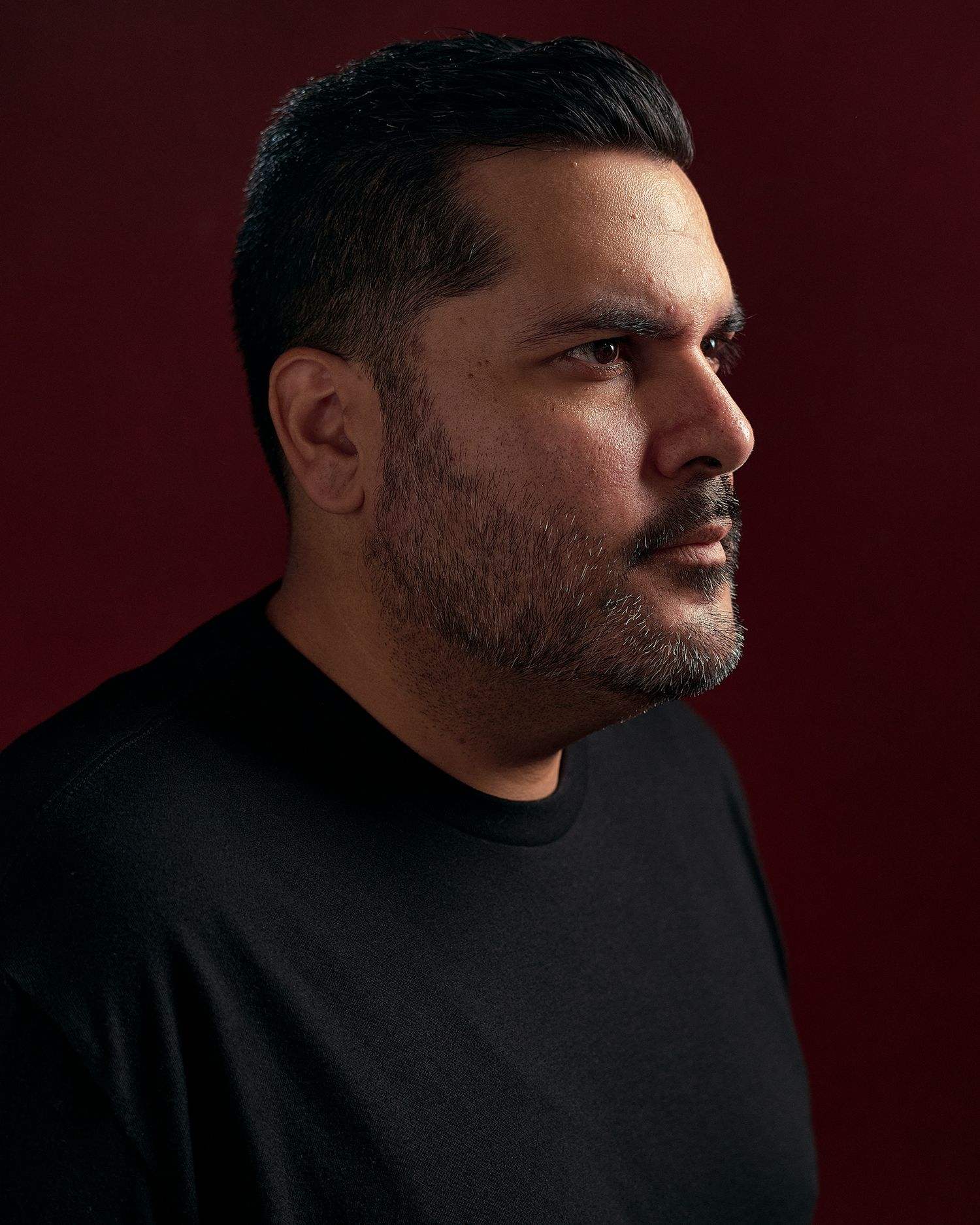 A bearded man in a black sweater poses in profile against a dark red background in a dramatic portrait.
