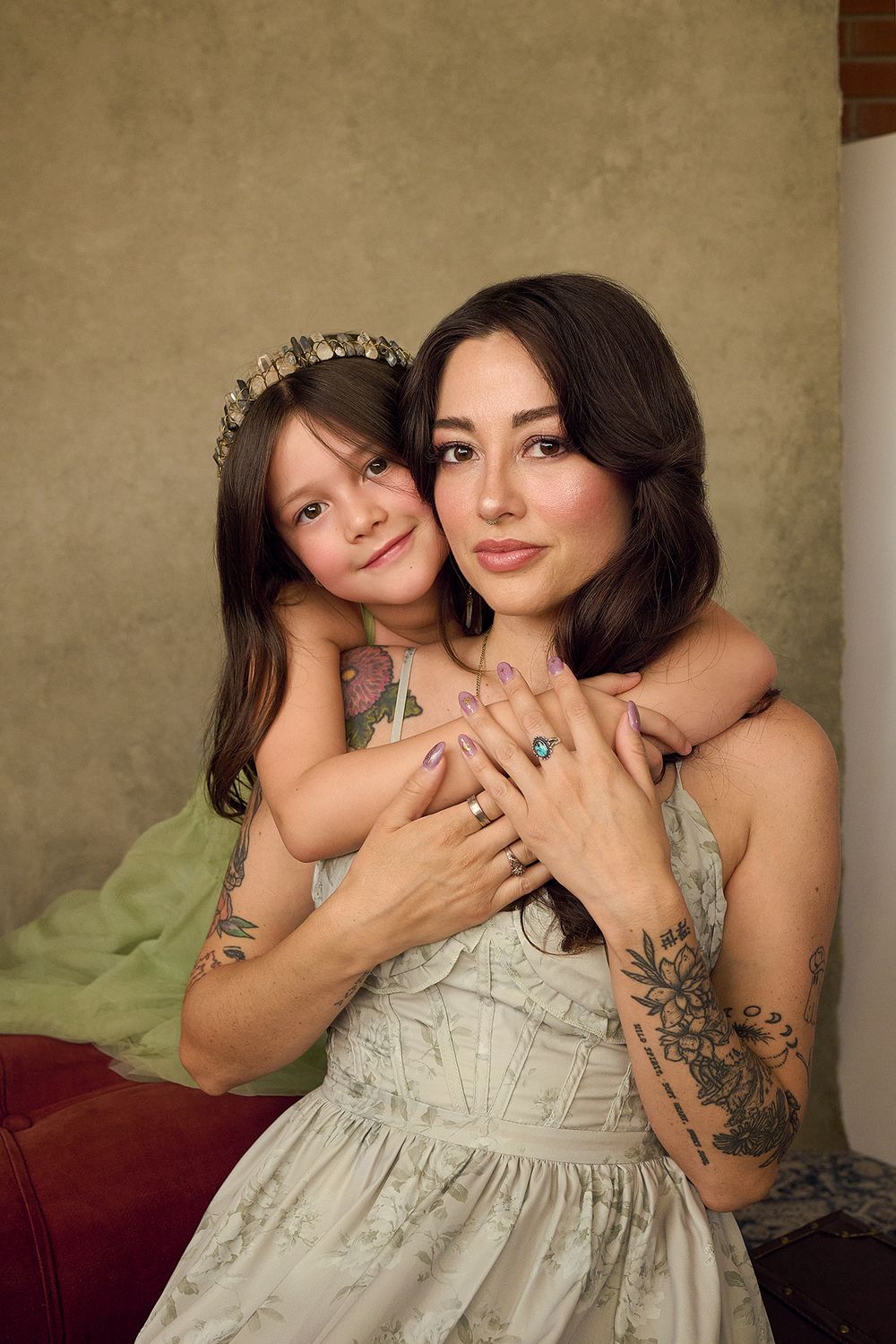 A young girl hugs a tattooed woman in a light dress, both smiling warmly in a cozy indoor setting.