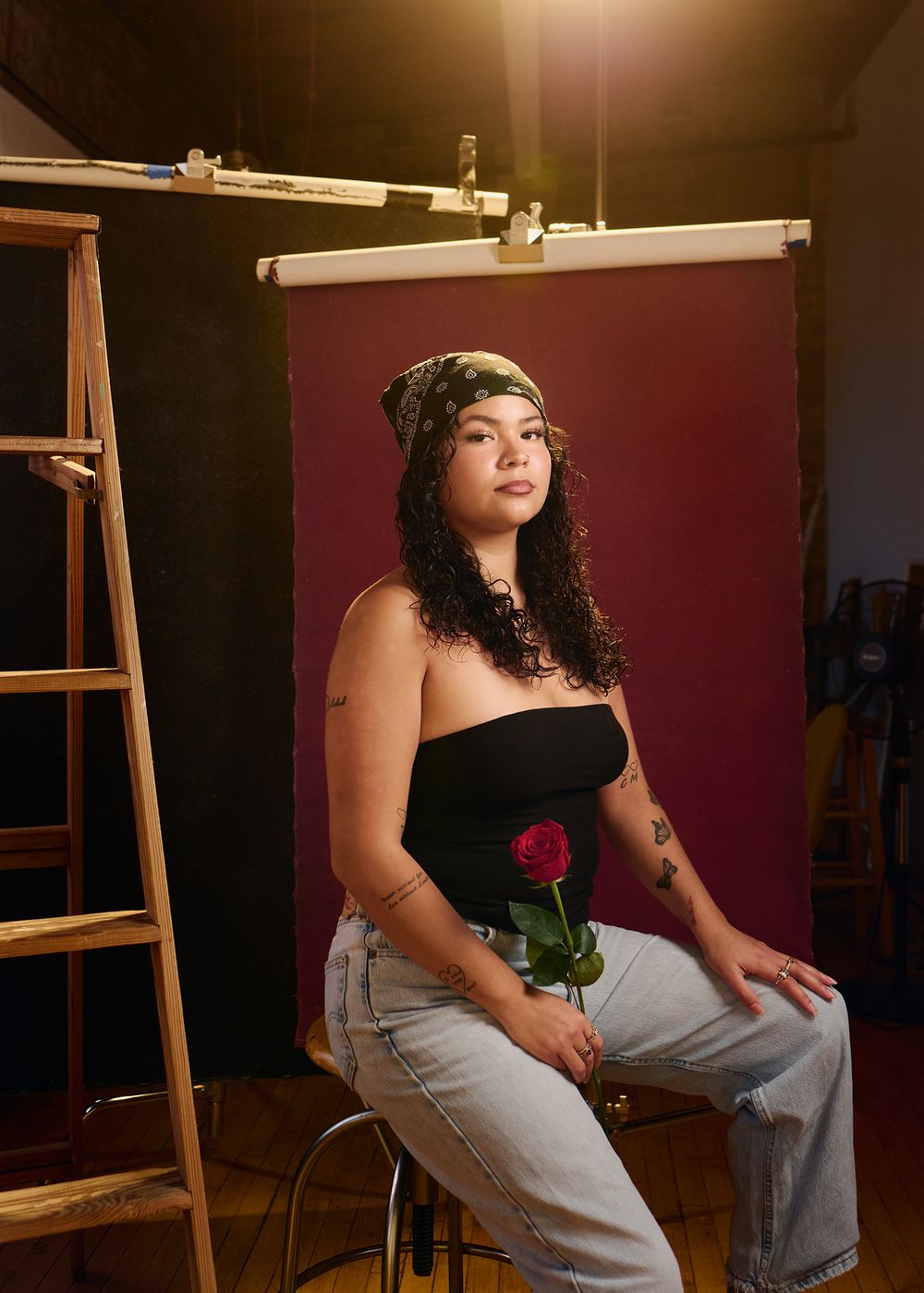 Woman in black tube top holding red rose seated on stool in front of burgundy backdrop in studio.