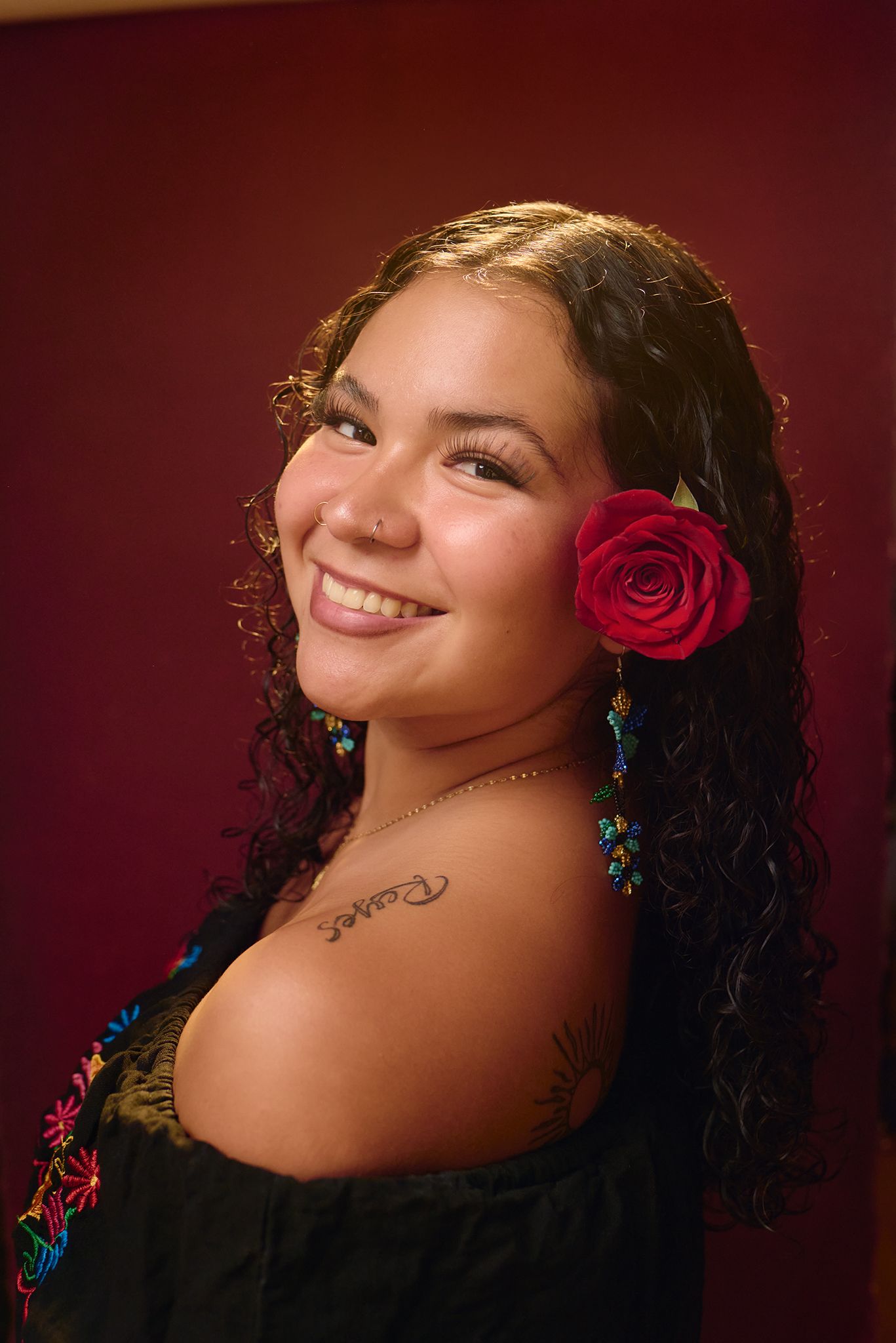 Smiling young woman with red rose in curly hair wearing off-shoulder embroidered top with shoulder tattoo.