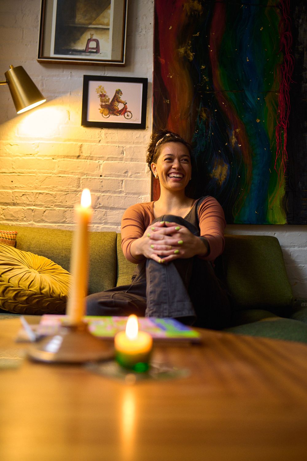 Joyful woman sitting on green couch laughing in cozy candlelit room with colorful wall art.