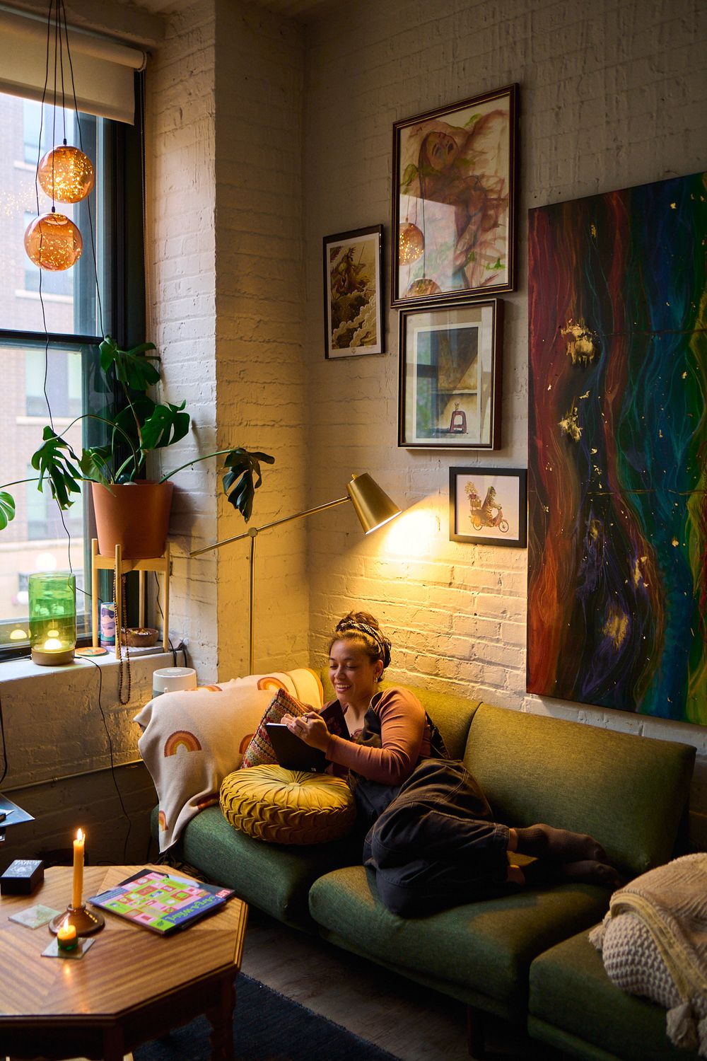 Woman relaxing on green sofa reading in warmly lit bohemian apartment with plants and artwork.
