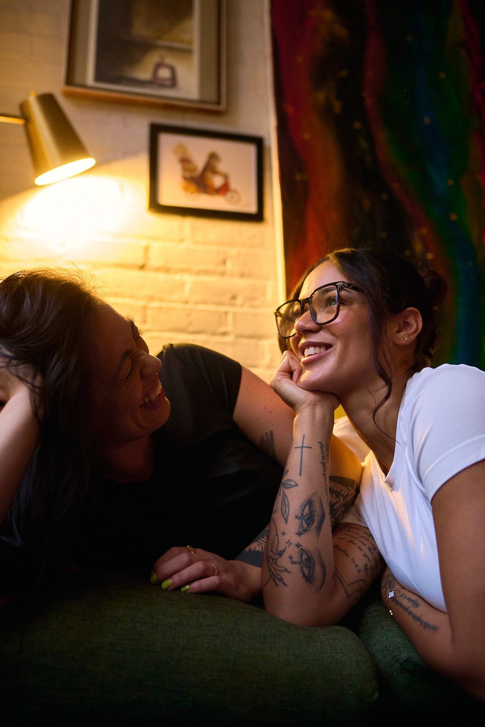 Two women smiling and laughing together on a green sofa in a warmly lit cozy room.