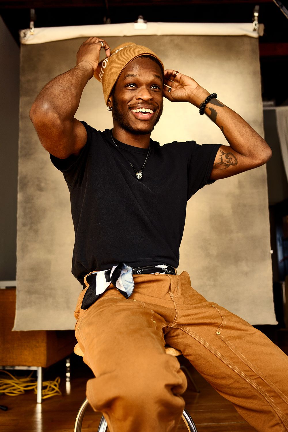 Smiling Black man in black t-shirt and tan cargo pants adjusting beanie hat in studio setting.