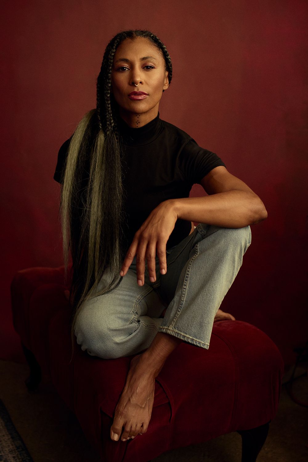 Woman with long braided hair sits on a red velvet stool in a black top and light jeans against a deep red backdrop.
