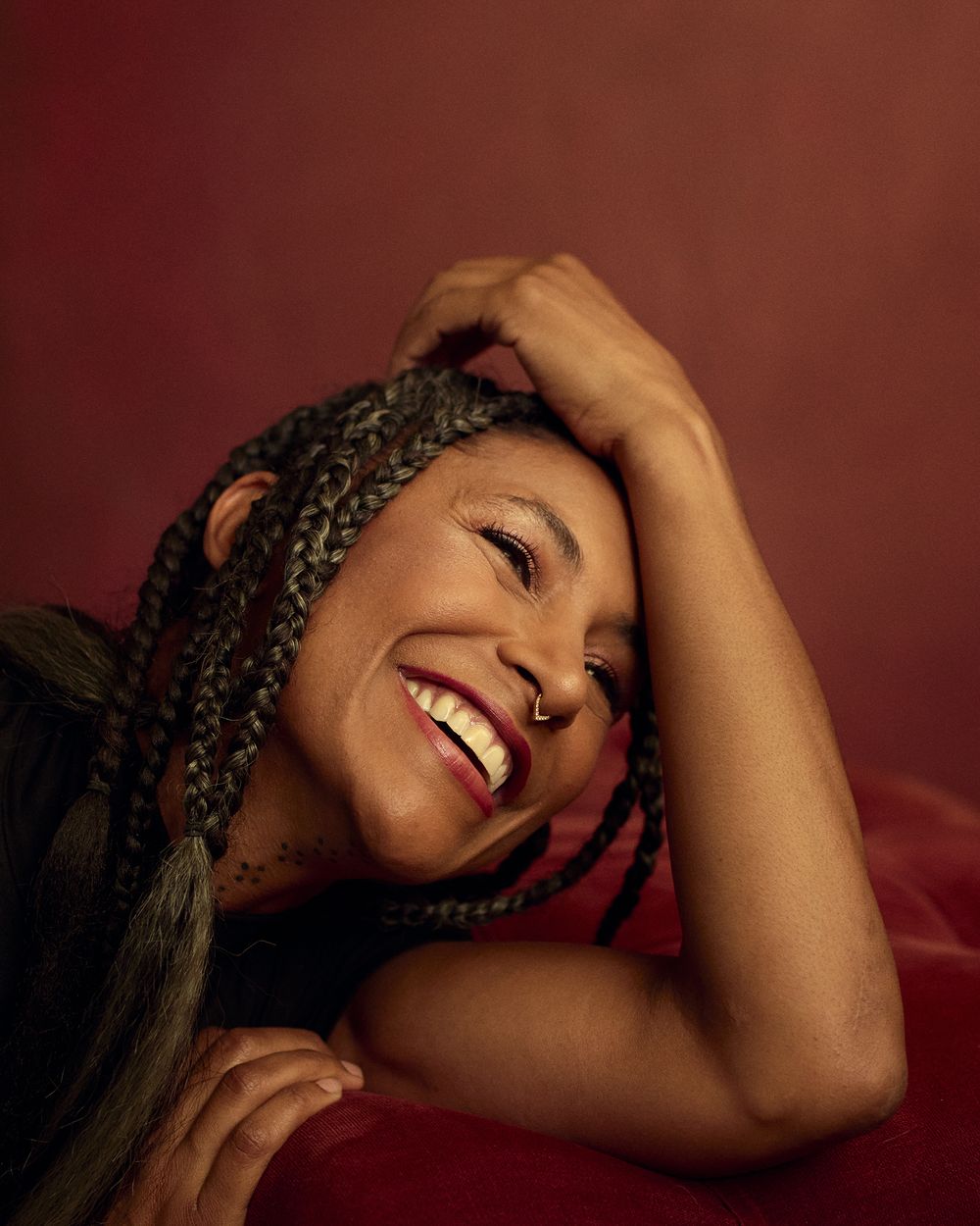 Smiling woman with long braids and a nose ring rests her head on her arm against a deep red background in a warm, joyful portrait.