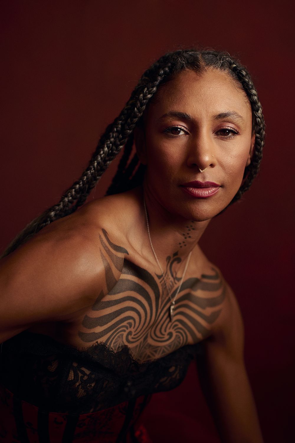 Woman with long braids and an intricate swirling chest tattoo wears a black lace corset and a nose ring against a deep red background.