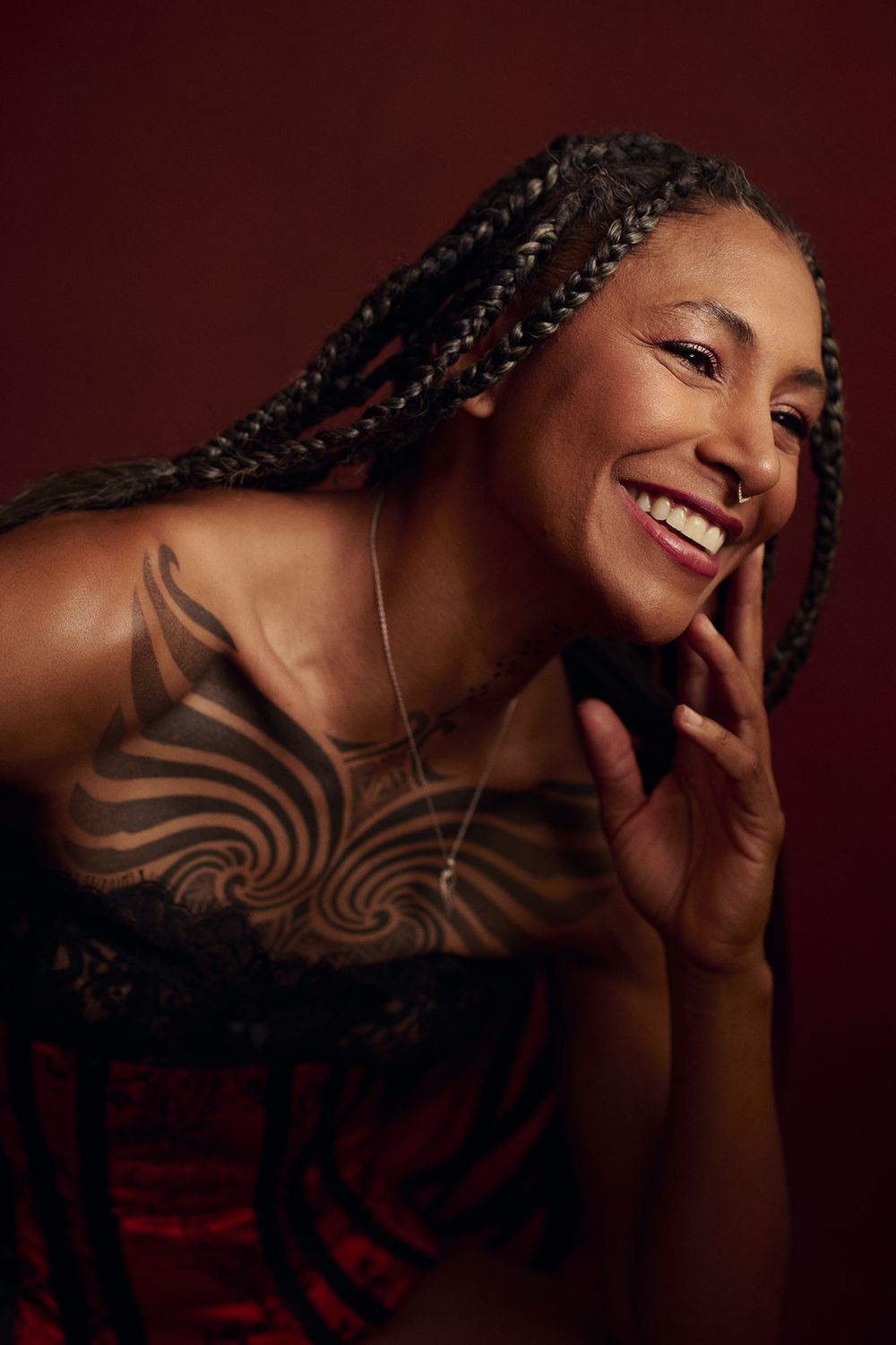 Smiling woman with long braids and a large chest tattoo leans forward, hand near her face, wearing a red corset against a dark red backdrop.