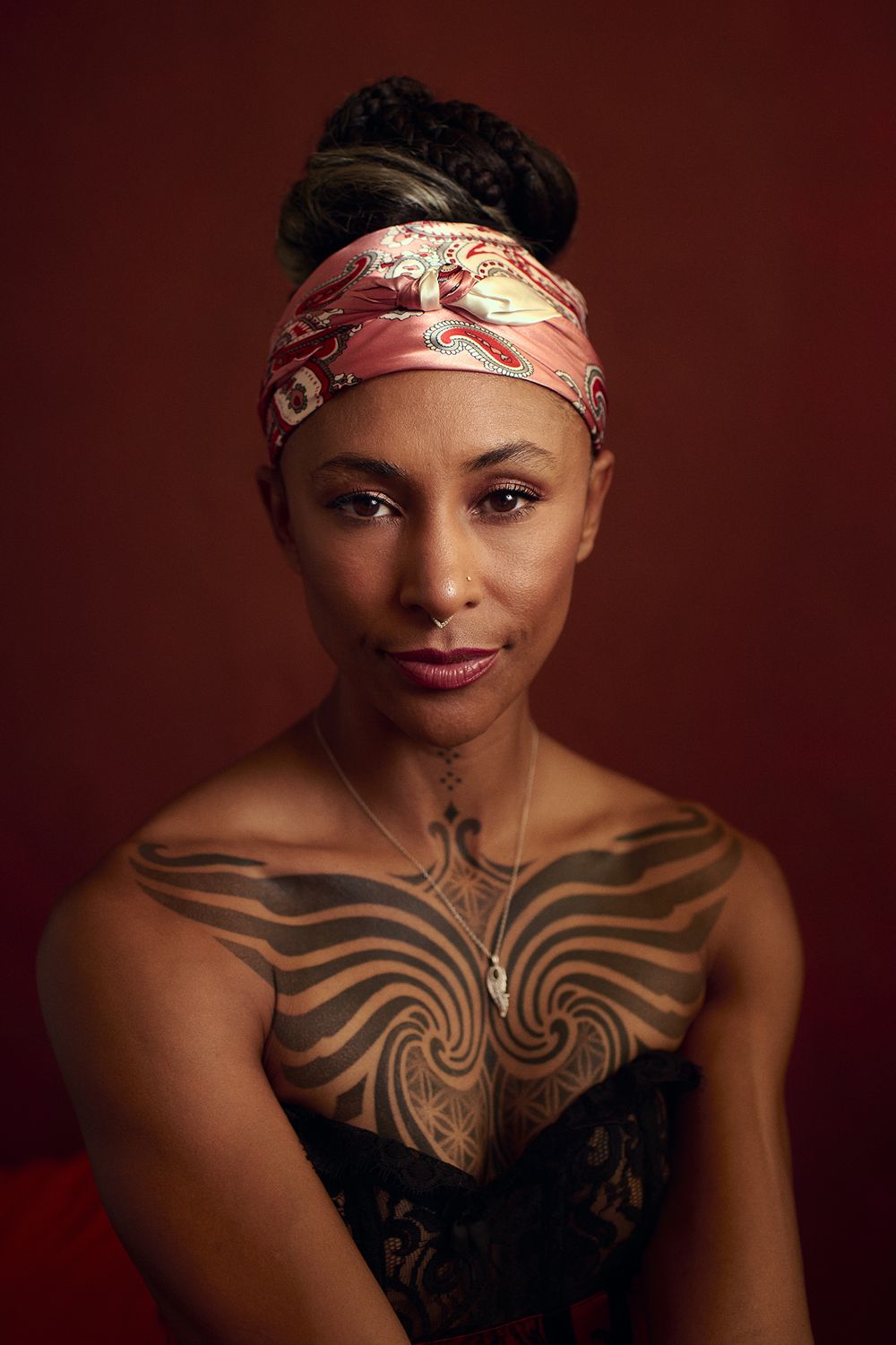 Portrait of a woman in a pink patterned headscarf with bold geometric chest tattoos and a pendant necklace against a red background.