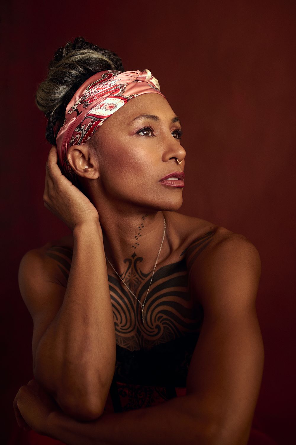 Woman with a pink patterned headband and tribal chest tattoos rests her head on her hand, gazing upward against a deep red backdrop.