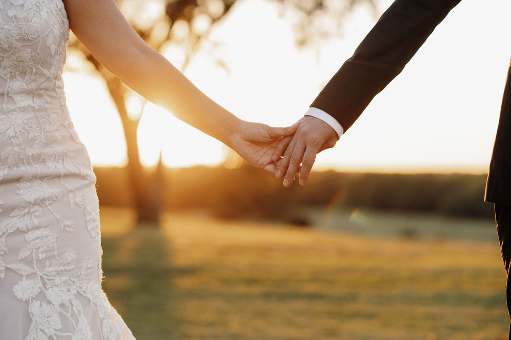 A couple holds hands during sunset in a romantic outdoor setting with golden light filtering through trees.