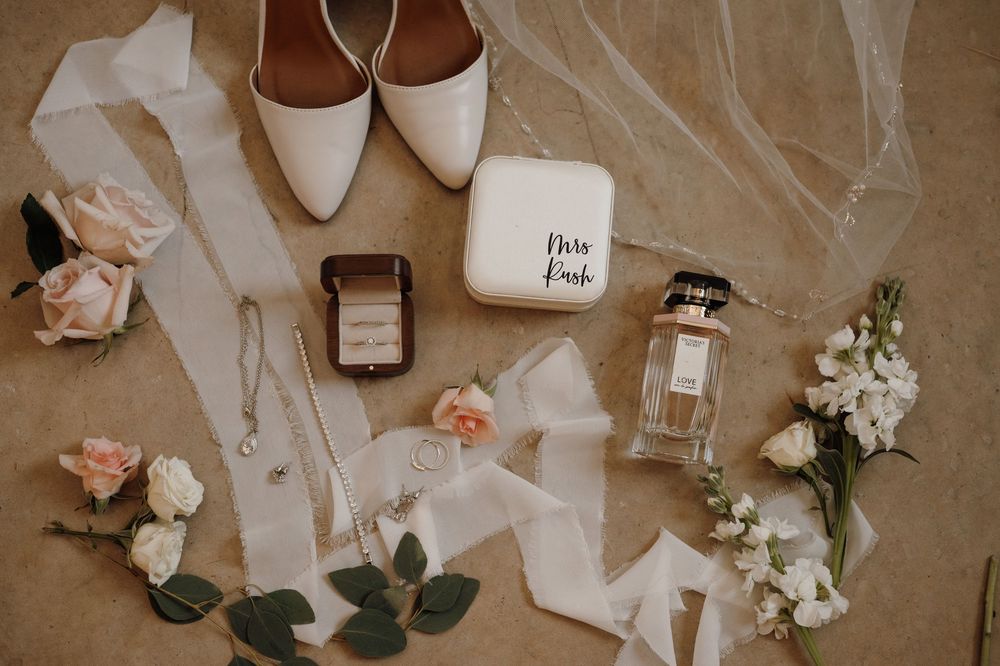 Wedding accessories including white shoes, perfume bottles, flowers and ribbons arranged on beige background.
