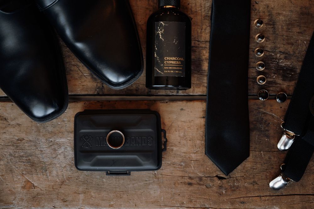 A black leather necktie lies flat on a rustic wooden surface next to a leather wallet and other accessories.