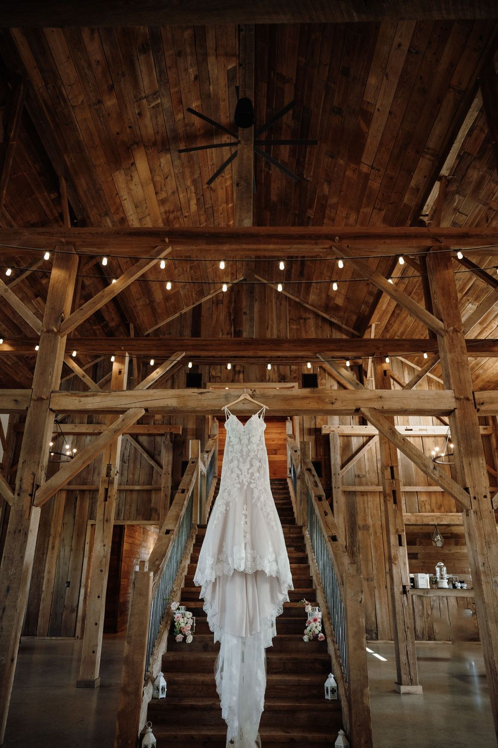 Wedding dress hanging in rustic wooden barn with exposed beams and string lights creating romantic ambiance.