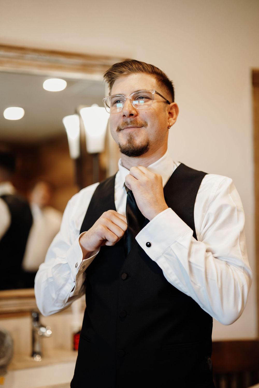 A person adjusts their tie while wearing a black vest and white dress shirt in a well-lit room.