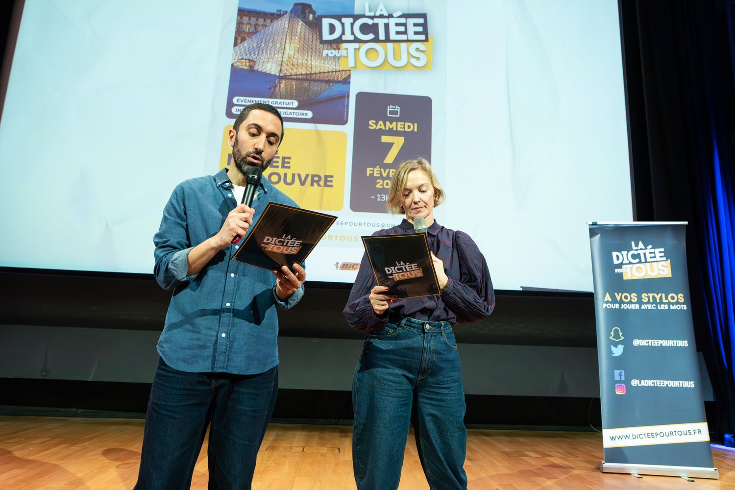 Two presenters in matching blue blazers share the stage at a business conference or event.