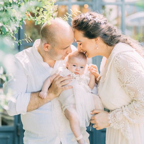 An intimate family portrait outdoors with a baby in white, surrounded by spring greenery.