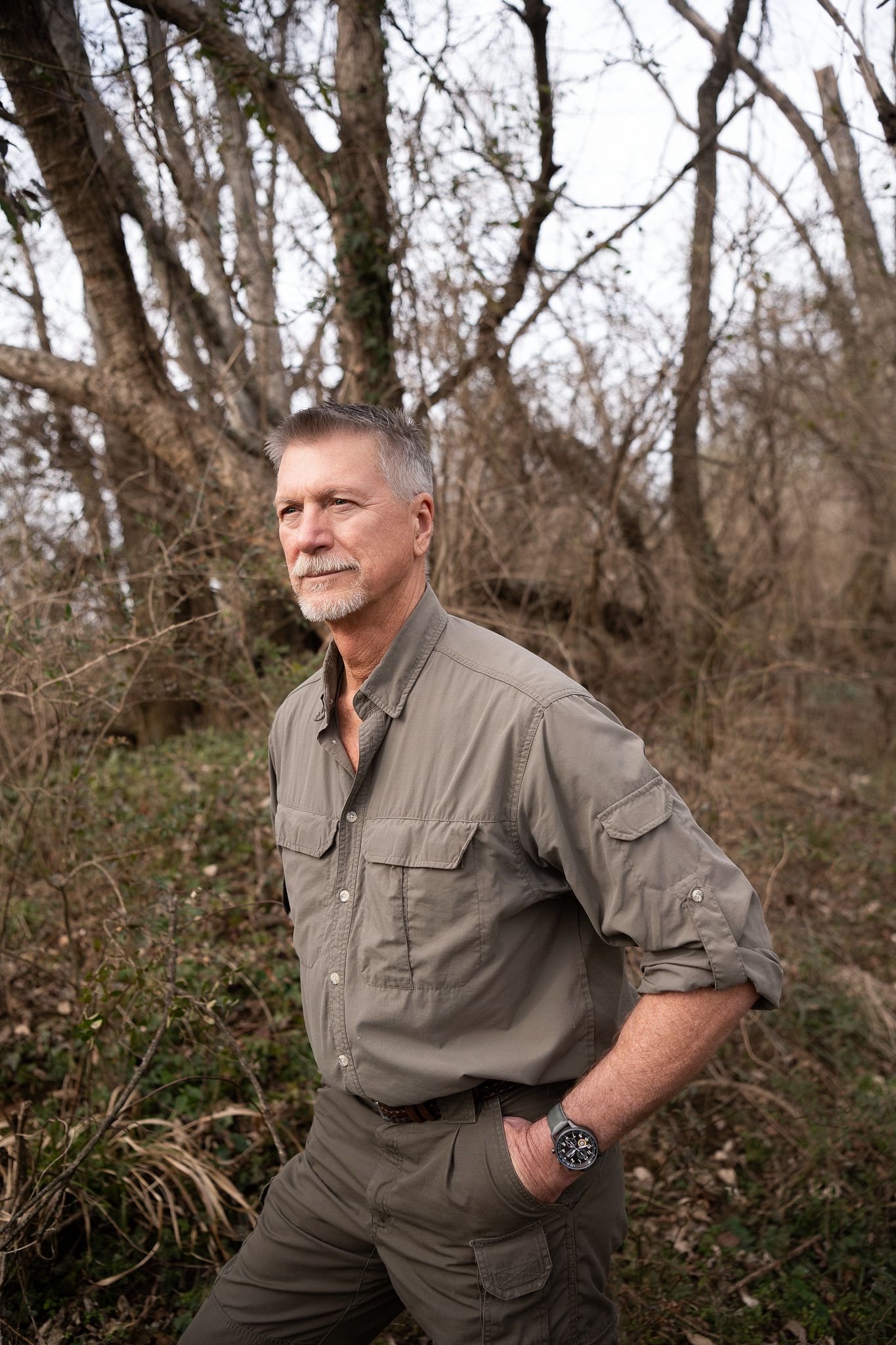 Person wearing tan outdoor clothing stands in front of wooded background in three similar poses.