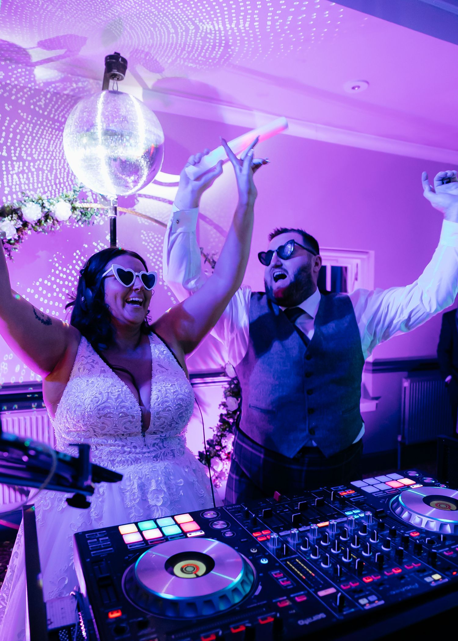 Wedding couple celebrating at DJ booth with disco ball and purple lighting at reception party.