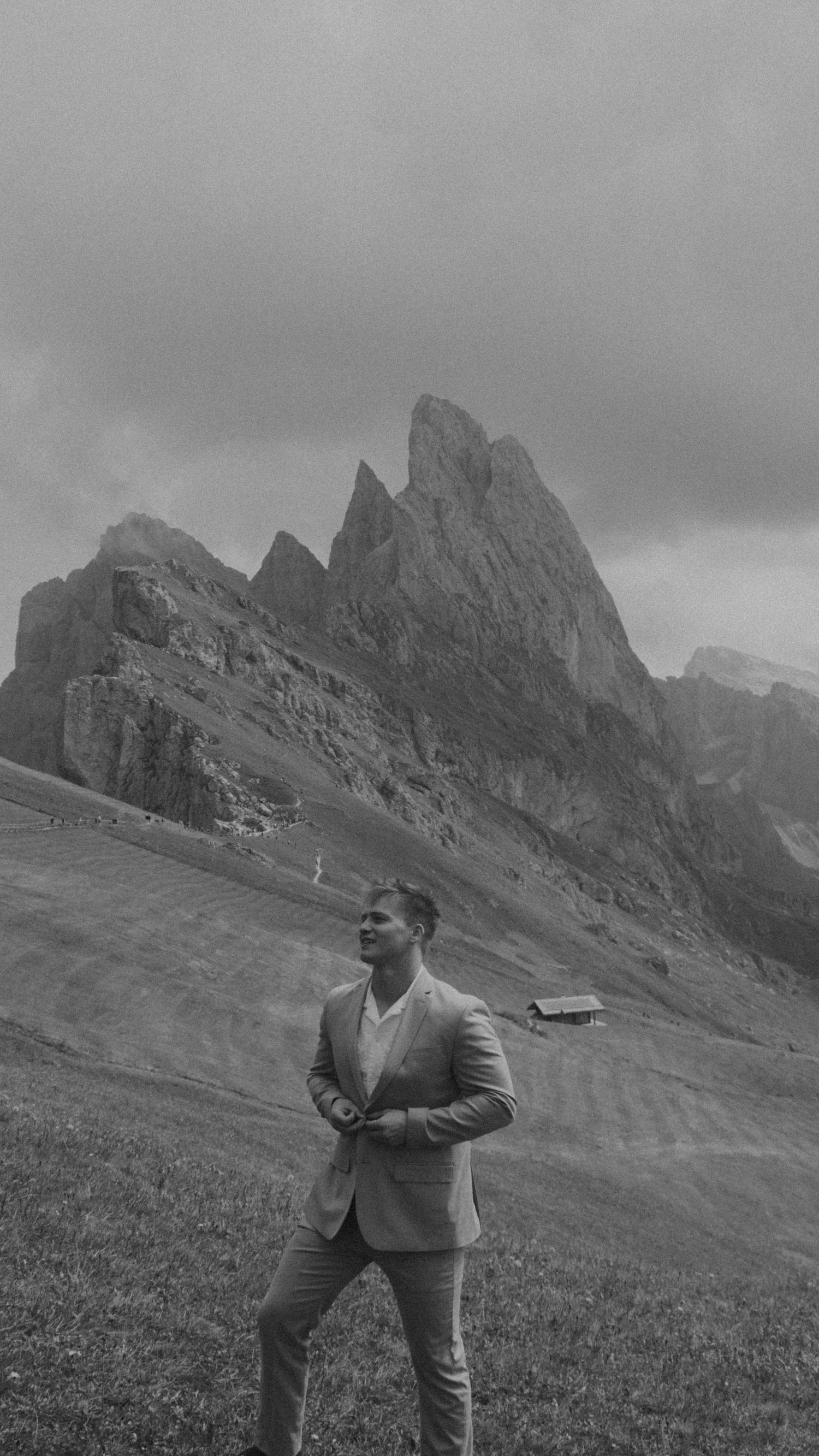 A person in a suit stands on a grassy slope with dramatic Dolomite mountain peaks rising behind them in black and white.