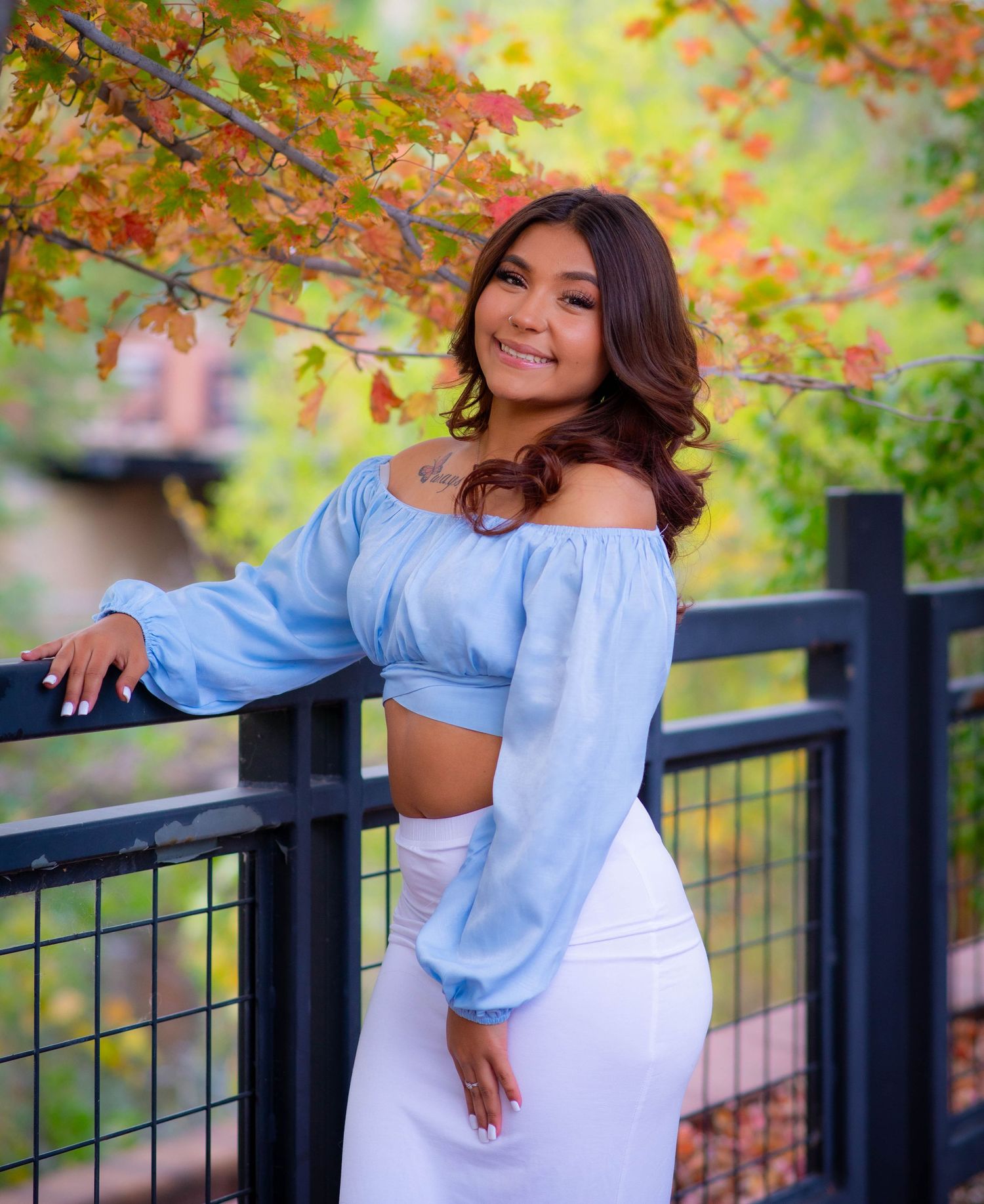 Woman in blue crop top and white skirt posing by railing with autumn leaves in background.