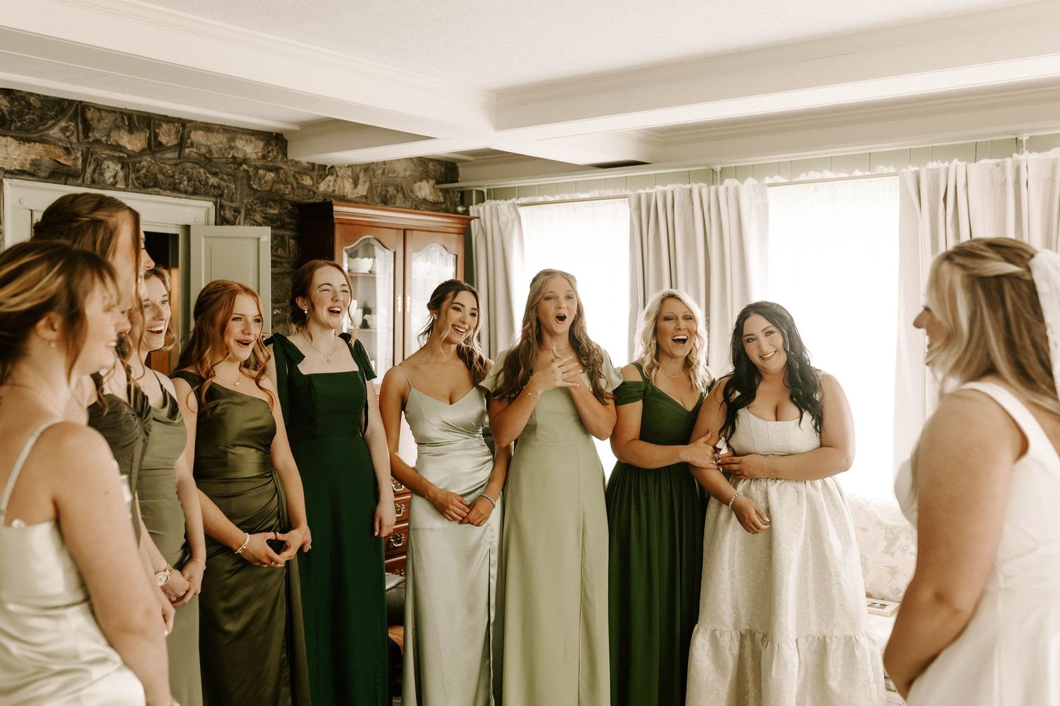 Bridesmaids in green and neutral dresses reacting joyfully to bride in white gown indoors.