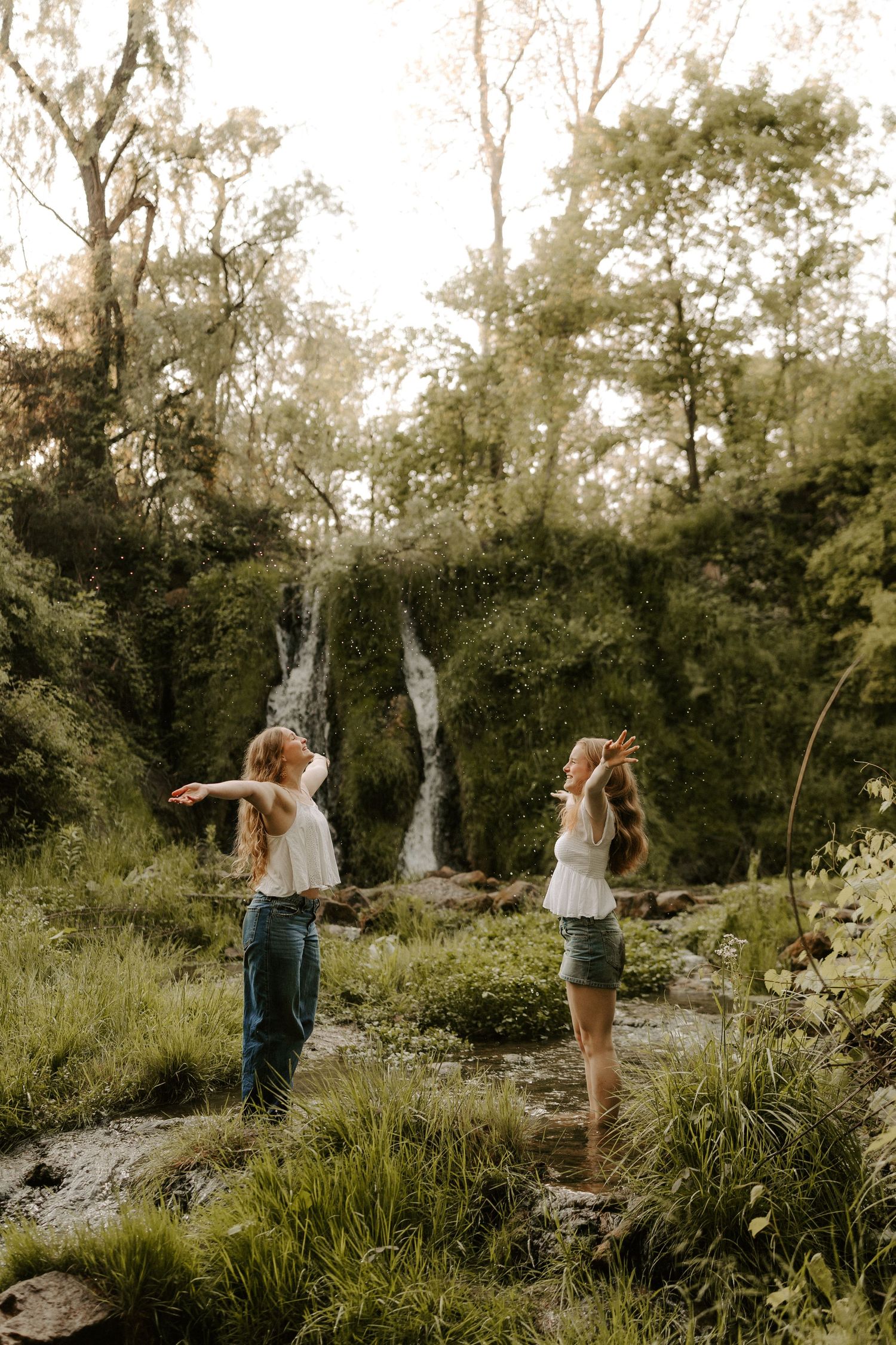 Two women in white tops pose playfully near waterfall in lush forest.