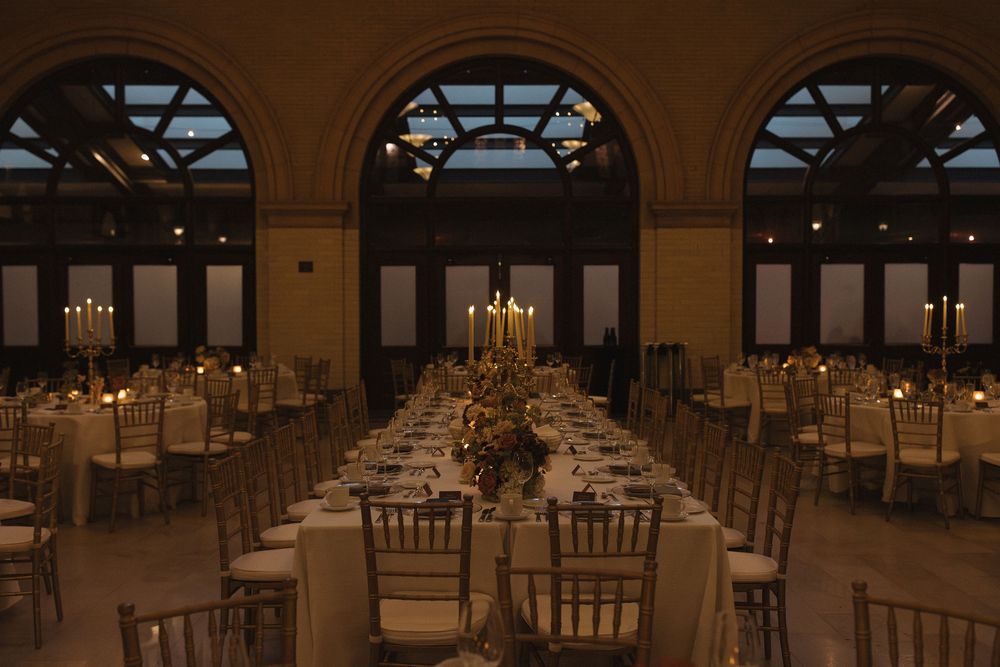 Elegant wedding reception hall with long tables, chiavari chairs, and arched windows illuminated by warm candlelight.