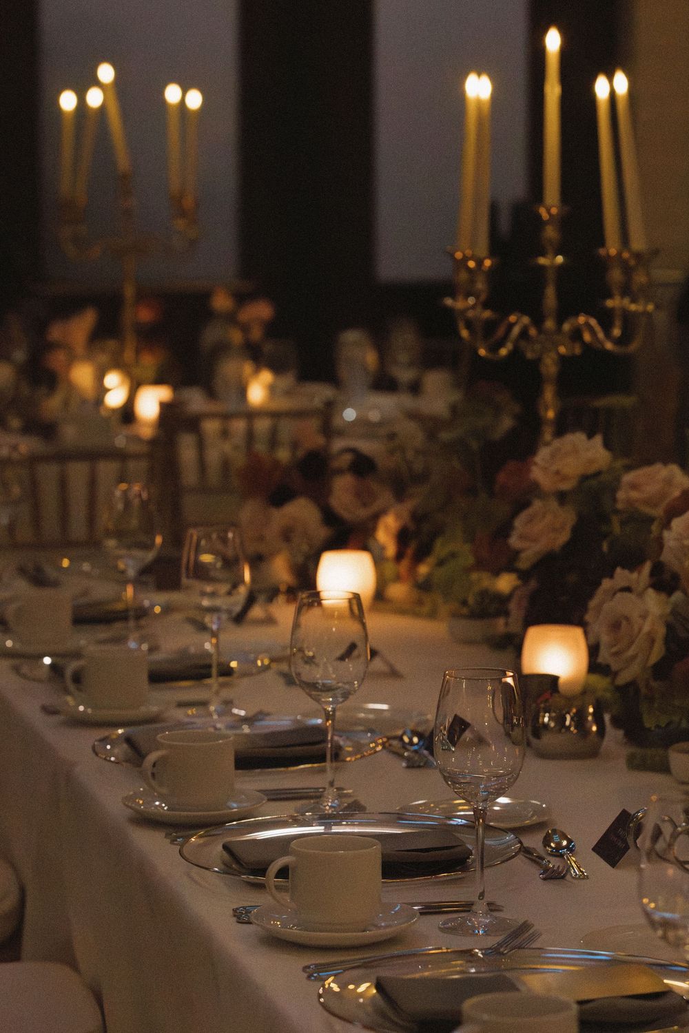 Elegant candlelit dinner table set with white linens, glowing tapers in brass holders, and delicate place settings.