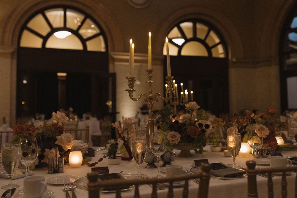 Elegant wedding reception tables set with candles and floral centerpieces in a dimly lit ballroom with arched windows.