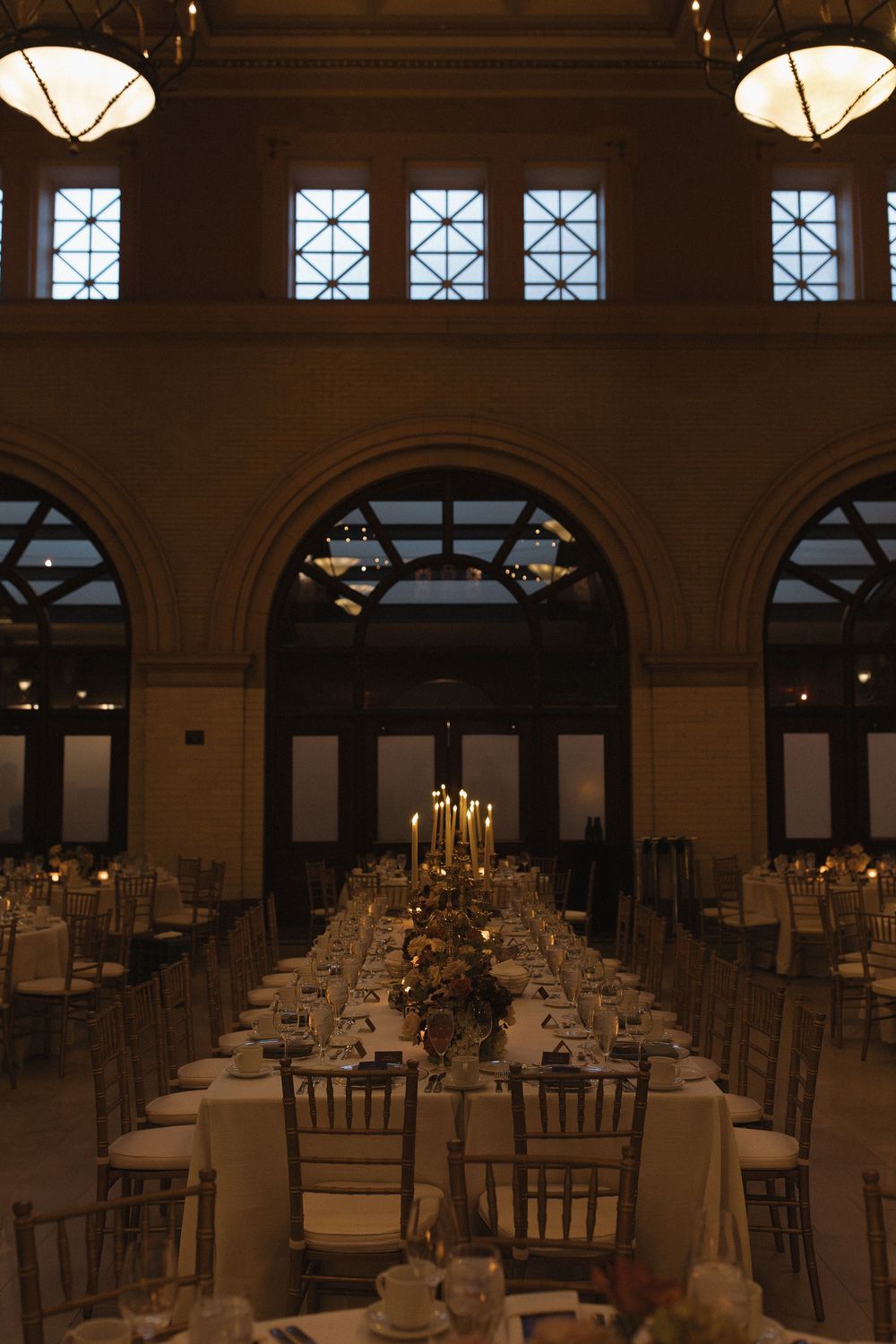 Elegant wedding reception venue with multiple tables arranged in rows under arched windows and crystal chandeliers.