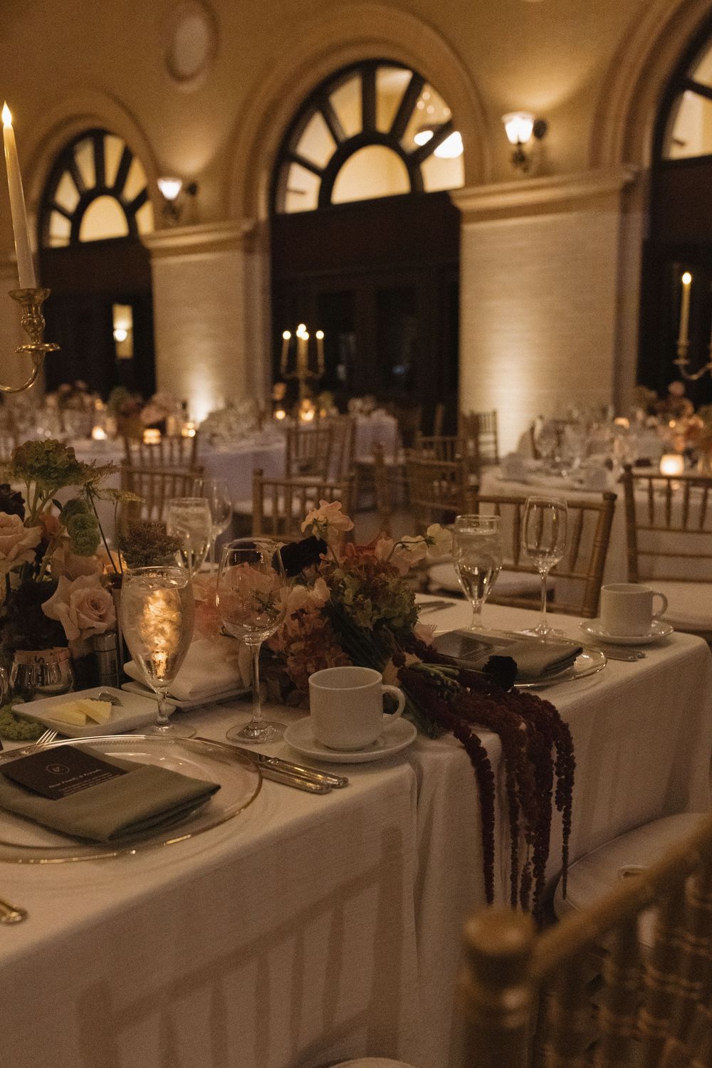 Formal dining tables set with white linens and elegant place settings under arched windows in a grand ballroom.