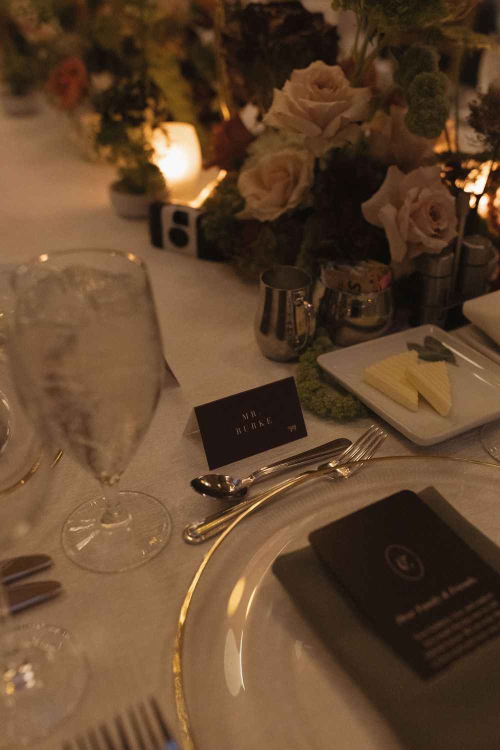 Elegant table setting with white linens, gold cutlery, and dark menu cards in warm candlelight.
