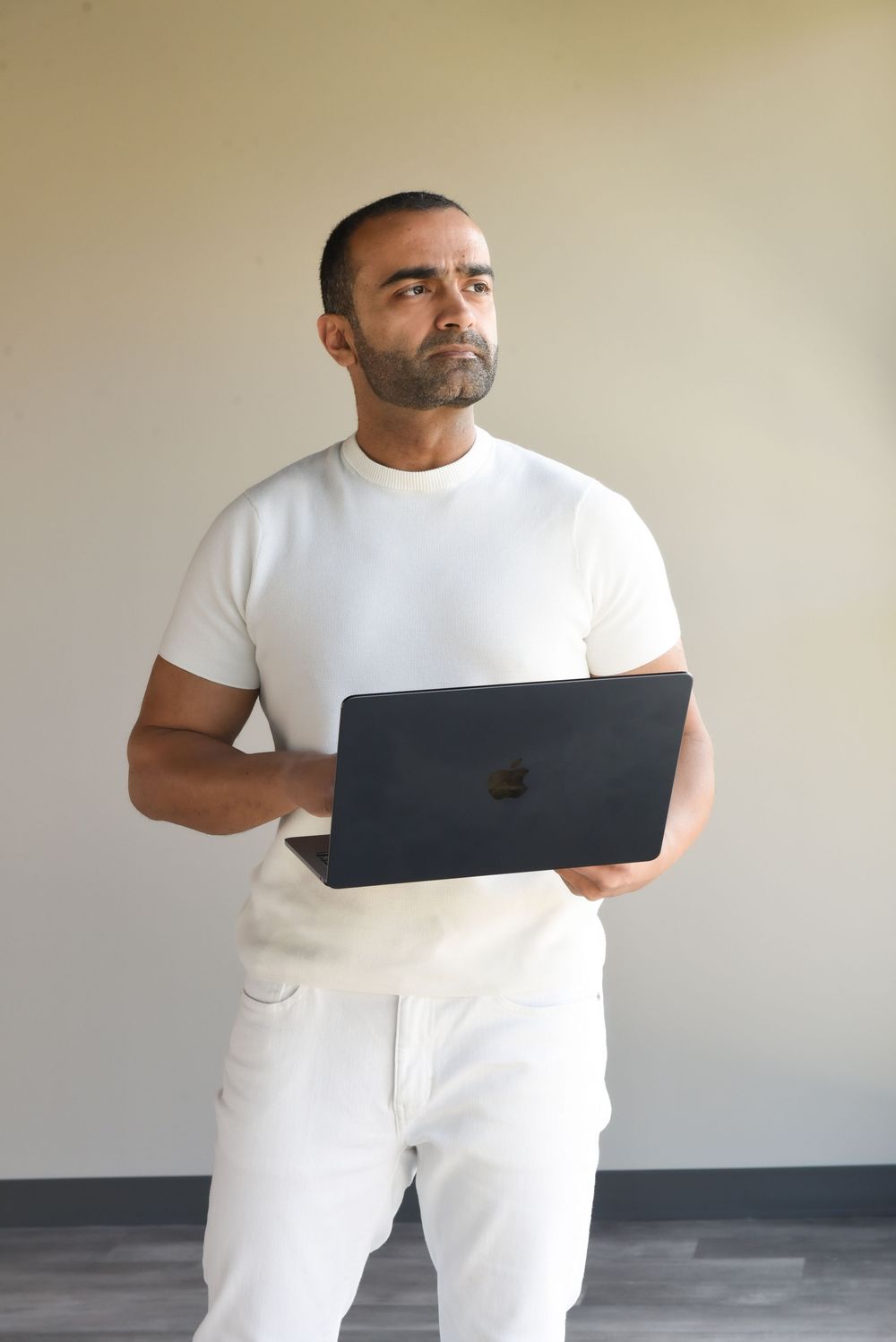 A person in white clothing holds a black laptop while standing against a light-colored wall.