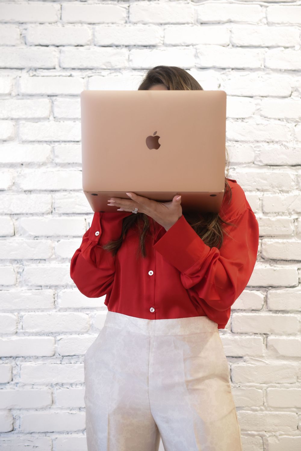 Someone in a red button-up shirt and white pants holds a rose gold MacBook in front of their face against a white brick wall.