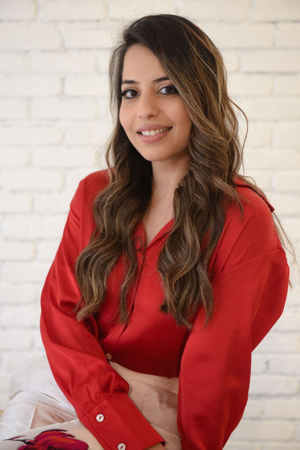 A person in a bright red blazer sits against a white brick wall with a warm smile and flowing brown hair.