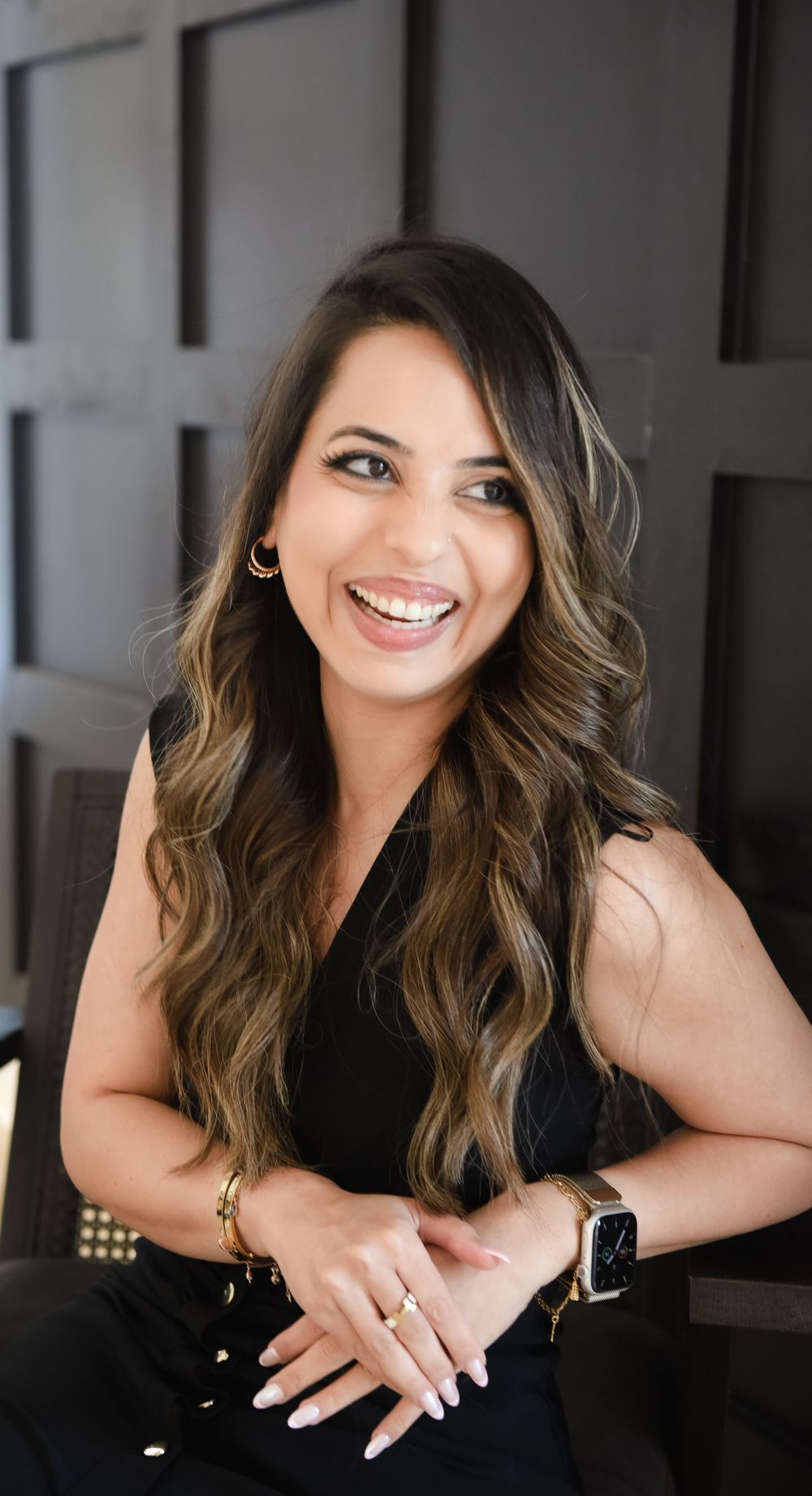 Professional headshot of someone with long wavy brown ombre hair wearing a black sleeveless top and smiling warmly.
