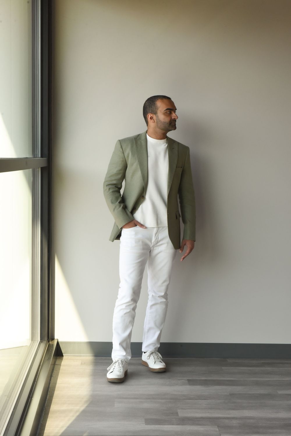 Four sequential fashion photos showing a stylish outfit with white pants, sage green blazer and white sneakers against grey wall.