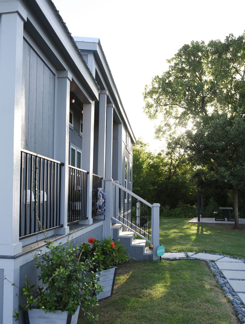 Modern gray mobile home exterior with black metal railings, potted flowers, and landscaped yard in late afternoon light.
