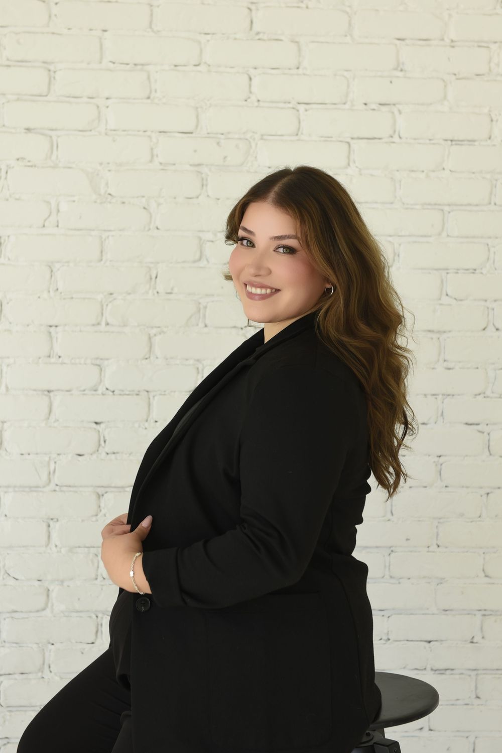 Plus size model in black blazer poses confidently against white brick wall in professional photoshoot series.