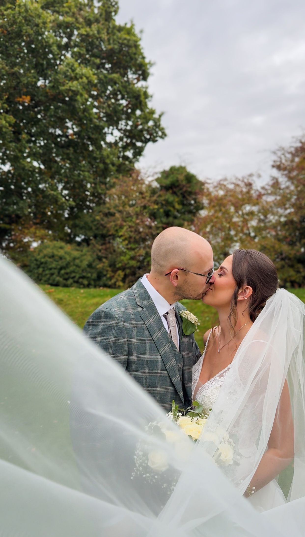 A romantic wedding couple shares a tender kiss outdoors with flowing veil and autumn trees in the background.