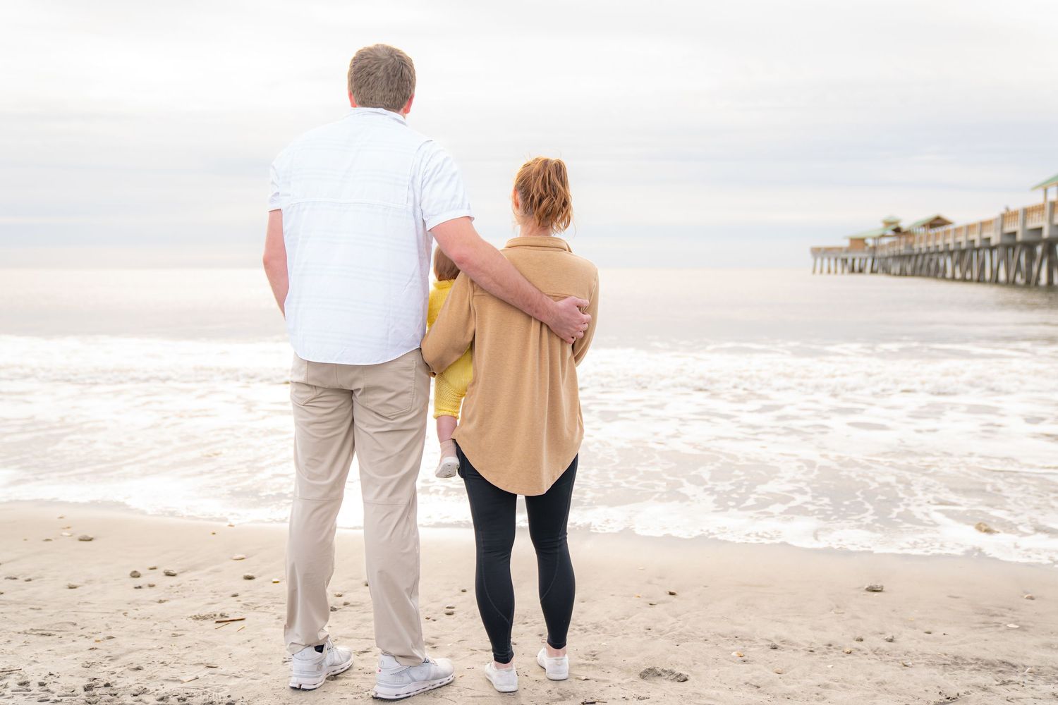 Two people walk hand in hand along a sandy beach with a wooden pier visible in the distance.