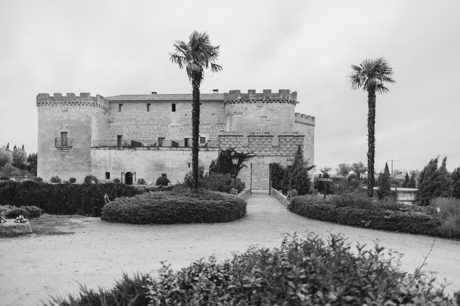 Black and white photograph of a historic Italian castle with two palm trees and manicured gardens in the foreground.