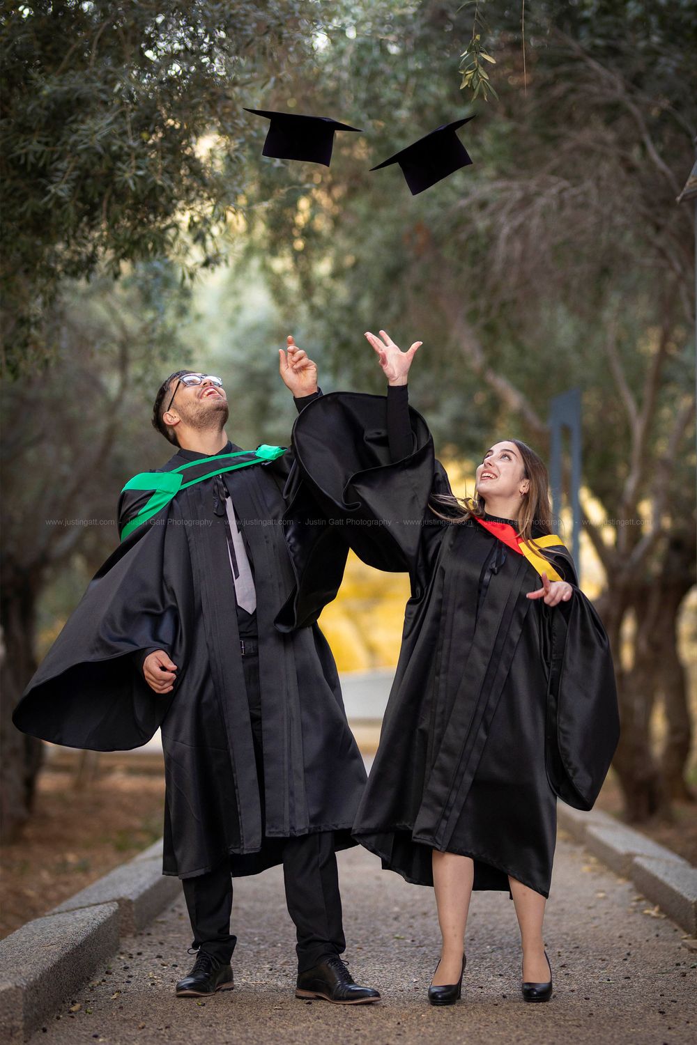 Graduates in black caps and gowns celebrating and tossing their caps on a tree-lined path.