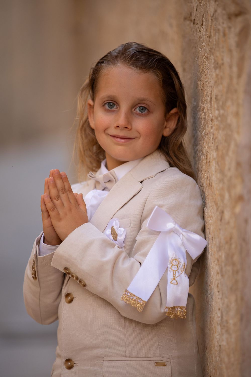 Child in formal white attire standing against stone wall with clasped hands.