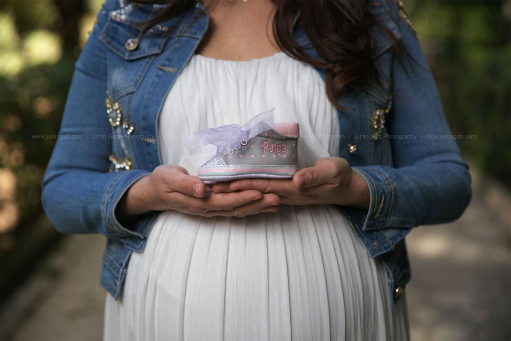 Pregnant belly in white dress with hands holding tiny pink baby shoes.