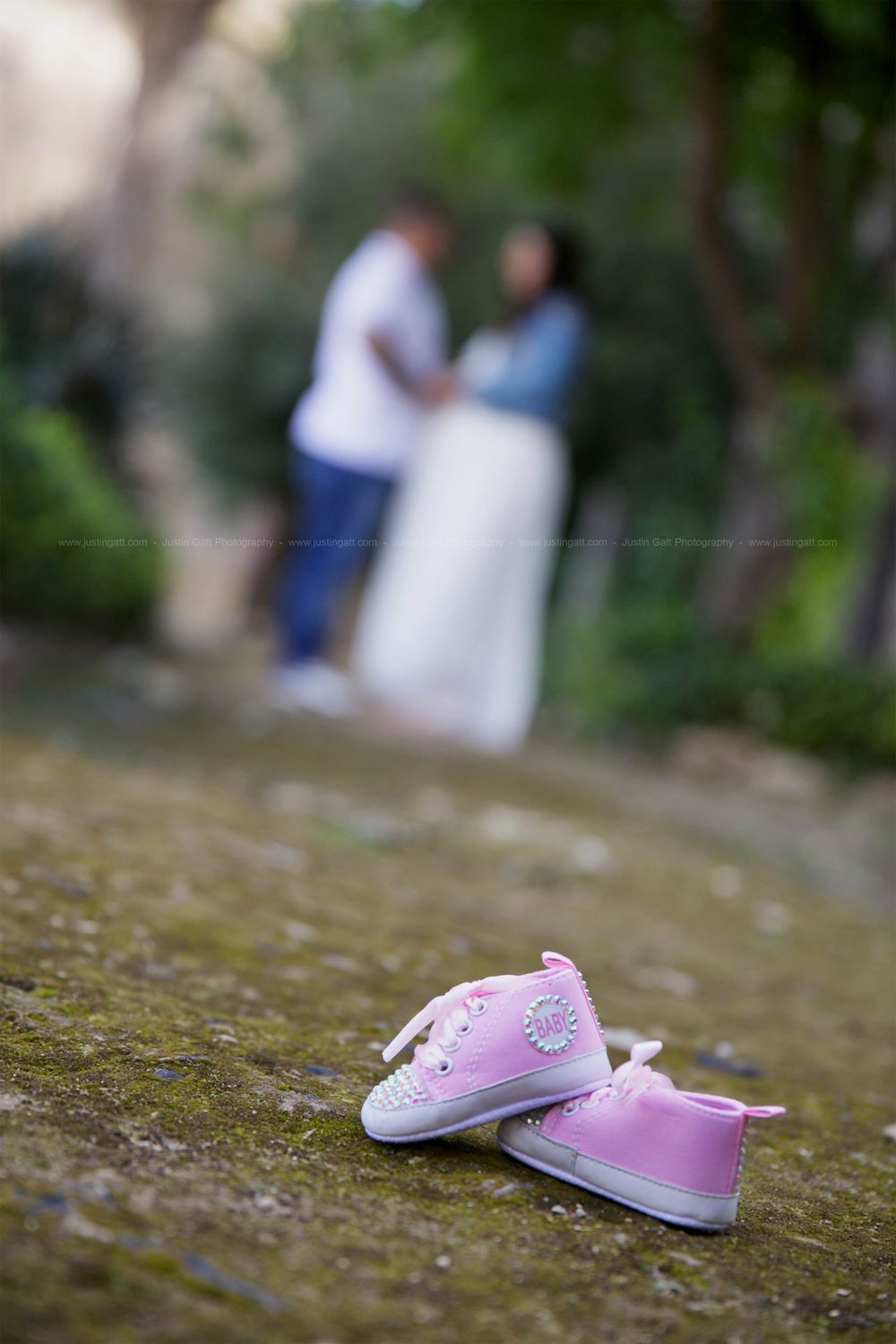 Pink baby shoes in foreground with blurred couple standing in garden background.