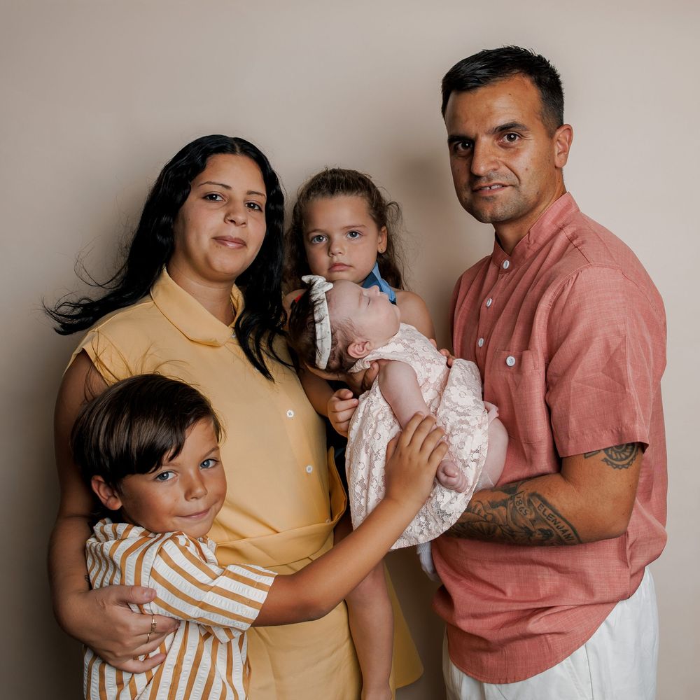 Family portrait of parents with three young children, including a newborn baby, posing together against a neutral wall.