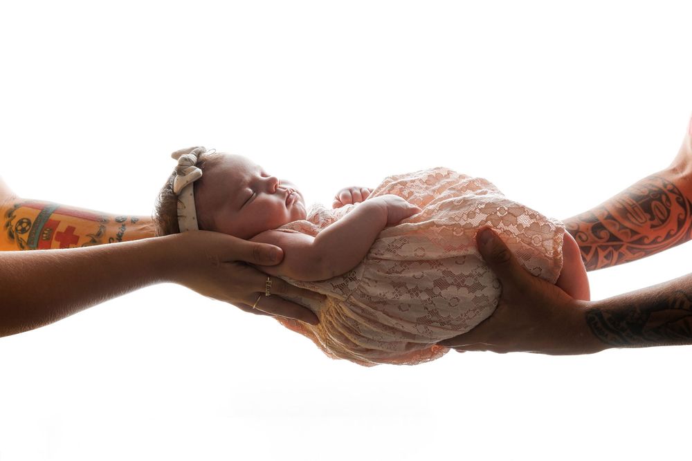 Two tattooed parents cradle their sleeping newborn baby wrapped in lace on a white background.
